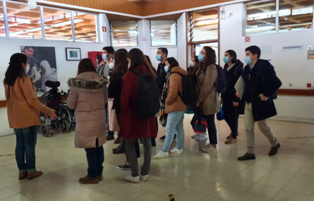 Grupo de pessoas em visita a espaço interior. Todas usam máscara de proteção individual. Ao fundo, na parede, alguns quadros. Vê-se ainda uma cadeira de rodas.
