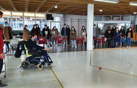 Em primeiro plano uma pessoa em cadeira de rodas a praticar boccia e a lançar a bola. Ao fundo, a assistir, mais de uma dezena de pessoas. Todas usam máscara de proteção individual.