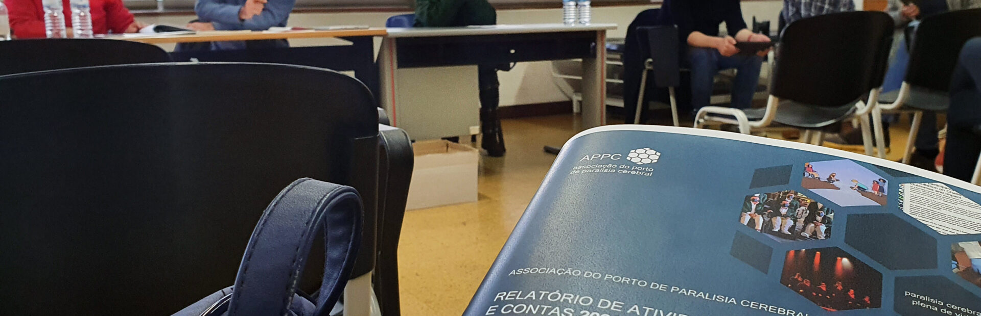 Em primeiro plano uma versão impressa do Relatório de Atividades e Contas 2021 da APPC. Mais ao fundo, desficadas, três pessoas sentadas junto de uma mesa. Ao lado outras três pessoas (igualmente sentadas).