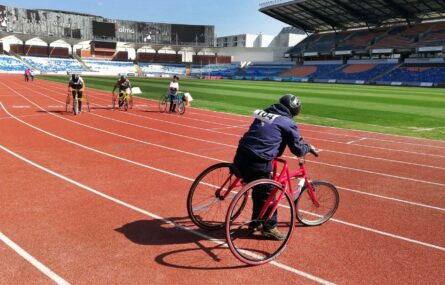 Cinco praticantes de tricicleta em pista. Relvado mais ao fundo e bancadas a toda a volta.