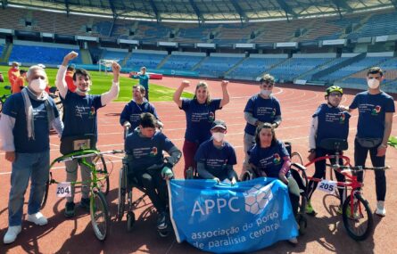 Fotografia de uma dezena pessoas em pista de estádio. Junto destas pessoas está uma bandeira azul da APPC. Ao fundo as bancadas. Em plano intermédio outras pessoas em atividade e a serem fotografadas.