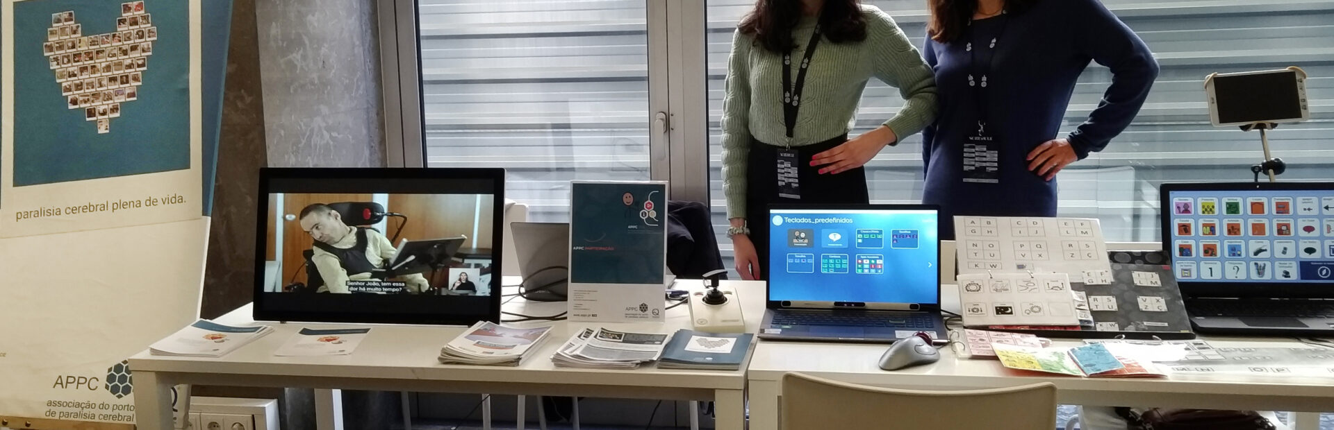 Duas mesas com diverso material informático, papéis e panfletos pousados. Atrás das mesas duas pessoas, em pé. À esquerda um quadro da Associação do Porto de Paralisia Cerebral. São ainda visíveis várias cadeiras e uma caixa de plástico pousada no chão.