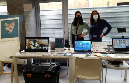 Duas mesas com diverso material informático, papéis e panfletos pousados. Atrás das mesas duas pessoas, em pé. À esquerda um quadro da Associação do Porto de Paralisia Cerebral. São ainda visíveis várias cadeiras e uma caixa de plástico pousada no chão.