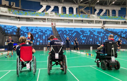 De costas, em cadeira de rodas, três elementos da seleção nacional de boccia. Mais ao fundo outros elementos de várias seleções. Em panorama as bancadas do pavilhão ainda sem público.