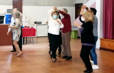 Três pares em plena dança. Ao fundo, junto da parede, algum material e uma mesa com cadeiras.