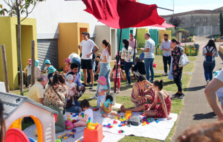 Fotografia de espaço ajardinado exterior. Cerca de duas dezenas de pessoas convivem e conversam. Crianças e adultos aparentam divertir-se. Os rostos das crianças foram todos desfocados.