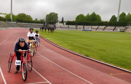 Três praticantes de tricicleta alinhados, em pista. À direita da imagem relvado com várias pessoas em corrida ou treino. Bancadas e árvores mais ao fundo.