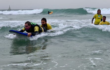 Pessoa deitada em prancha a praticar, no mar, surf adaptado. Uma outra pessoa acompanha e apoia a prancha. Mais ao fundo outras duas pessoas sorriem.