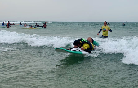 Pessoa deitada em prancha a praticar, no mar, surf adaptado. Uma outra pessoa acompanha e apoia a prancha. Mais ao fundo, também dentro de água, uma outra pessoa acompanha a atividade. Ao fundo da imagem mais pessoas dentro de água.