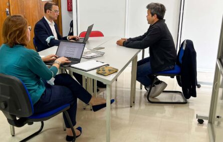 Duas mesas juntas. À volta da mesa estão três pessoas sentadas, Duas das pessoas utilizam computadores portáteis enquanto a terceira pessoa está, aparentemente, a falar.