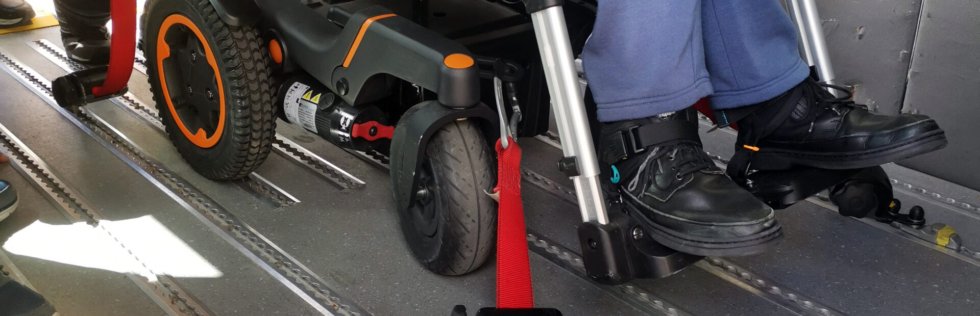 Fotografia de parte inferior de uma cadeira de rodas – notando-se a forma como está a ser fixada ao piso de uma viatura.