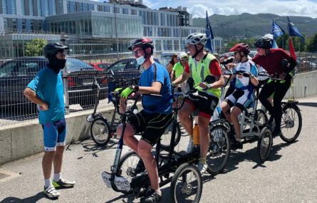 Quatro pessoas testam, par a par, duas bicicletas adaptadas. Ao lado estão mais bicicletas adaptadas e algumas pessoas a assistir. Ao fundo da imagem são visíveis edifícios.