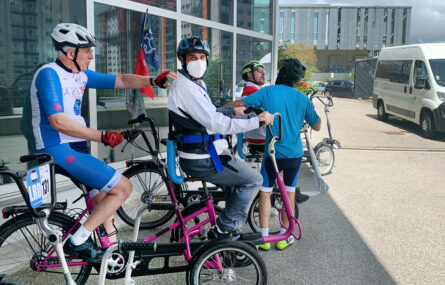 Quatro pessoas junto de três bicicletas adaptadas. Mais ao fundo da imagem estão algumas viaturas e vê-se ainda um edifício.