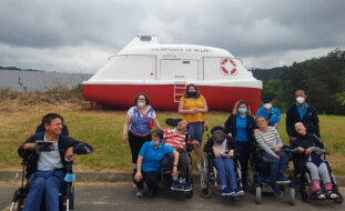 Grupo de 11 pessoas em espaço exterior. Atrás das pessoas, em zona relvada, está o que aparenta ser um pequeno barco (de apoio ou de socorro) que apresenta a inscrição “Voluntários de Melres”. Árvores ao fundo.