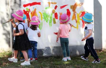Cinco crianças pintam um mural em parede. O mural já tem traços e desenhos de várias cores. Os rostos das crianças foram desfocados.