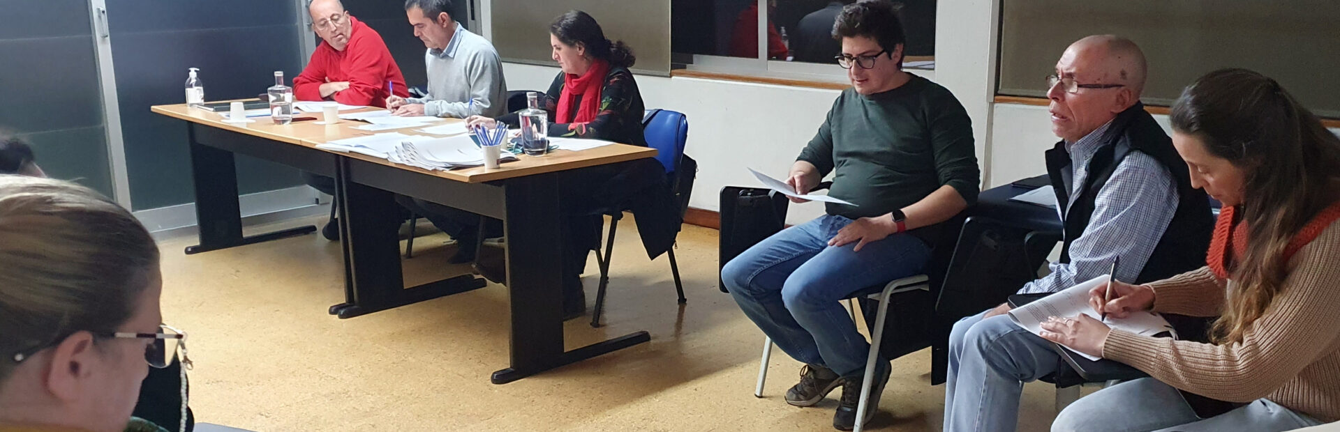 Fotografia de sala interior. Junto de duas mesas estão sentadas três pessoas. Ao lado outras três pessoas estão sentadas em cadeiras. Em primeiro plano, parcialmente visível, está uma outra pessoa a consultar algo no telemóvel