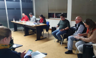 Fotografia de sala interior. Junto de duas mesas estão sentadas três pessoas. Ao lado outras três pessoas estão sentadas em cadeiras. Em primeiro plano, parcialmente visível, está uma outra pessoa a consultar algo no telemóvel