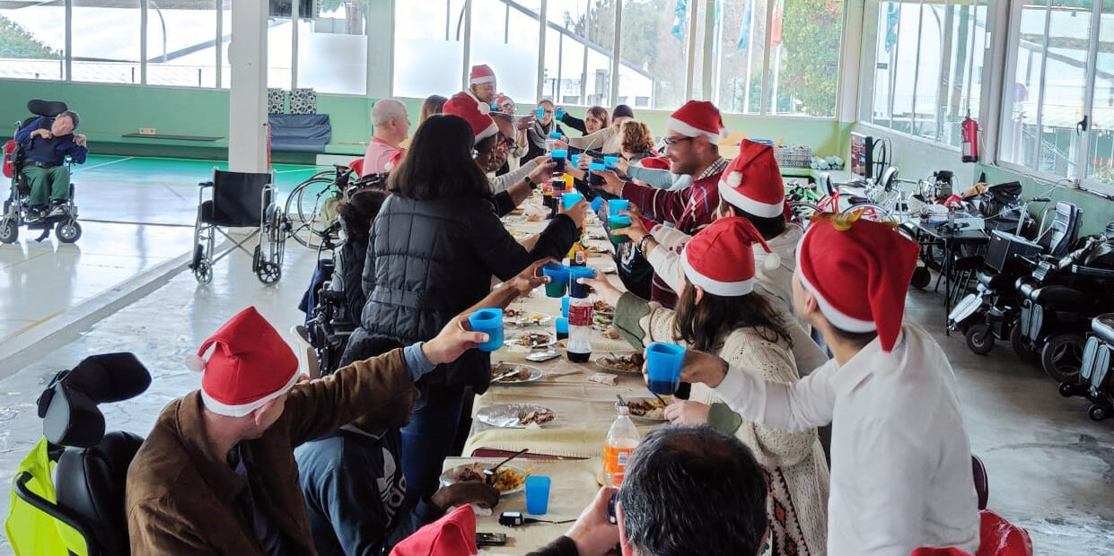 Mais de duas dezenas de pessoas sentadas (ou em pé) junto de uma mesa comprida. Estão a fazer um brinde segurando os copos nas mãos. Quase todas as pessoas estão a usar um gorro vermelho e alusivo ao Natal.