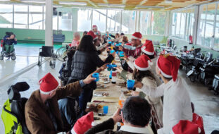 Mais de duas dezenas de pessoas sentadas (ou em pé) junto de uma mesa comprida. Estão a fazer um brinde segurando os copos nas mãos. Quase todas as pessoas estão a usar um gorro vermelho e alusivo ao Natal.