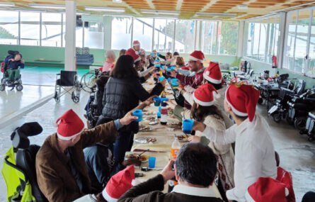 Mais de duas dezenas de pessoas sentadas (ou em pé) junto de uma mesa comprida. Estão a fazer um brinde segurando os copos nas mãos. Quase todas as pessoas estão a usar um gorro vermelho e alusivo ao Natal.