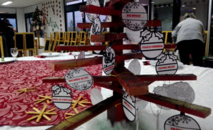 Fotografia a uma mesa com árvore de natal feita de pequenos pedaços de madeira. Na árvore estão penduradas bolas de Natal, impressas em papel, com vários nomes. Ao fundo da imagem estão algumas mesas, várias cadeiras e uma árvore de Natal.