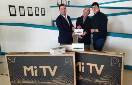 Três pessoas junto de equipamento eletrónico devidamente empacotado. Uma das pessoas entrega às outras uma pequena caixa.