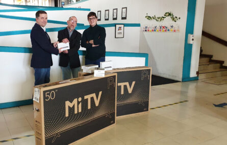 Três pessoas junto de equipamento eletrónico devidamente empacotado. Uma das pessoas entrega a outra uma pequena caixa. Ao fundo uma rampa de acesso e escadas.