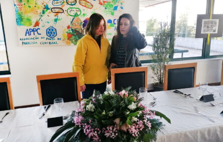 Duas pessoas junto de uma mesa – na qual está pousado, ao centro, uma ramo de flores.