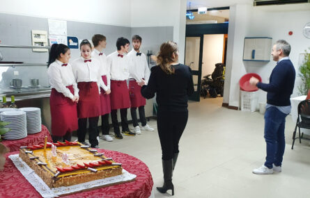 Em primeiro plano mesa com um bolo de aniversário. Mais ao fundo grupo de cinco jovens, trajadas com roupa para atendimento da refeição, ouvem indicações de dois adultos.
