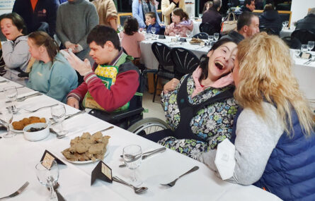 Grupo de pessoas, a conversar, à mesa. Além dos pratos, talheres e copos, já foram colocados alguns aperitivos. Uma das pessoas ri-se de forma evidente.