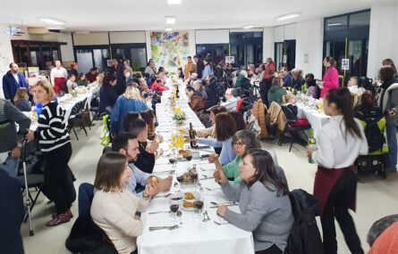 Perspetiva geral de três mesas, enormes e compridas, com perto de uma centena de pessoas. Além dos pratos, talheres e copos, já foram colocados alguns aperitivos, sumos e outras bebidas.