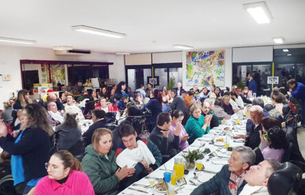 Perspetiva geral de três mesas, enormes e compridas, com perto de uma centena de pessoas. Além dos pratos, talheres e copos, já foram colocados alguns aperitivos, sumos e outras bebidas.