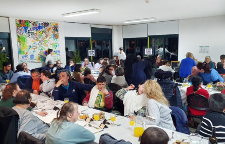 Grupo de pessoas, a conversar, à mesa. Além dos aperitivos, talheres, copos e sumos, já foram colocados pratos com sopa.