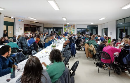 Ao fundo da imagem está um grupo de pessoas com vários instrumentos. Aparentam estar a interpretar uma música. Em primeiro plano, junto de uma mesa, quatro pessoas preparam-se para cortar o bolo de aniversário.