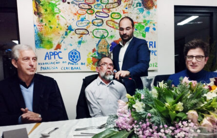 Plano aproximado de três pessoas sentadas à mesa. Em destaque está um ramo de flores. Uma quarta pessoa, de pé, segura um microfone para o elemento sentado ao centro que está a falar. Ao fundo, na parede, quadro colorido.