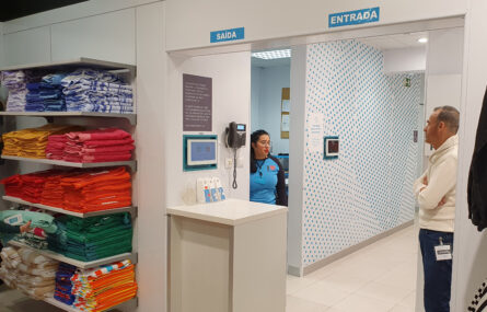 Imagem do interior de uma loja de roupa. À esquerda cinco prateleiras cheias de roupas e panos coloridos. Duas pessoas conversam junto da entrada de acesso à zona dos provadores de roupas.
