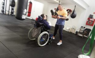 Pessoa em cadeira de rodas simula prática de boxe com o auxílio de uma outra pessoa, em pé, que segura acessórios desportivos.
