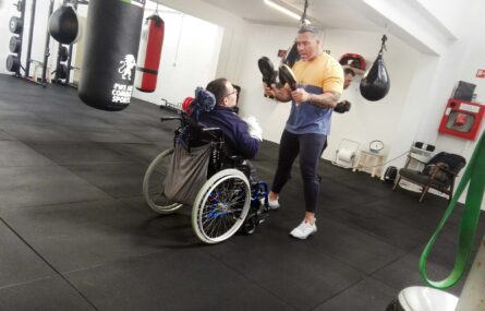 Pessoa em cadeira de rodas simula prática de boxe com o auxílio de uma outra pessoa, em pé, que segura acessórios desportivos.