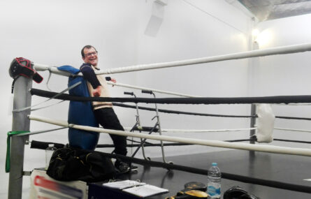 Pessoa sorridente apoiada às cordas dentro de ringue de boxe.