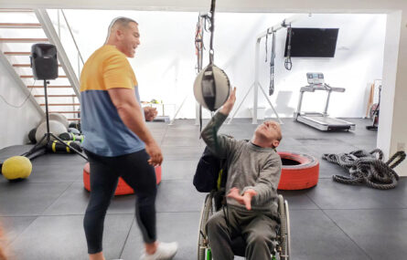 Pessoa em cadeira de rodas exercita-se com material de boxe. Ao lado outra pessoa assiste e incentiva. Diversos materiais estão espalhados no chão (mais ao fundo do ginásio).
