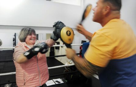 Pessoa simula prática de boxe com o auxílio de uma outra pessoa que segura acessórios desportivos.