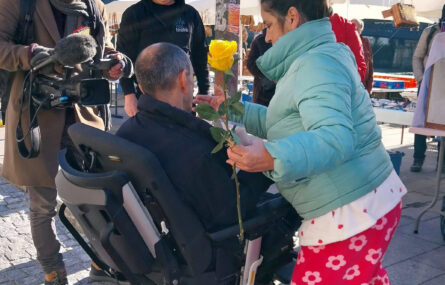 Pessoa em cadeira de rodas a tentar subir a um passeio sem acessibilidade. No lado direito está uma pessoa com uma rosa na mão. À esquerda um repórter de imagem (com câmara) e várias outras pessoas.
