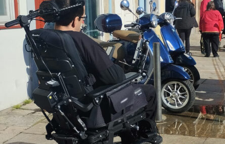 Pessoa em cadeira de rodas, num passeio, a constatar o facto de não conseguir transitar. À sua frente estão estacionadas, no passeio, várias motorizadas.