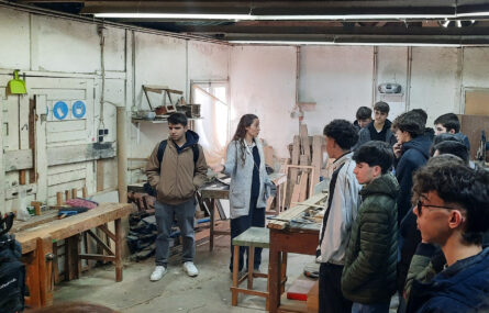 Fotografia de espaço interior no que aparenta ser uma marcenaria (com diverso material espalhado pelo chão e colocado em prateleiras nas paredes. Um grupo de alunos conversa com elemento da APPC.