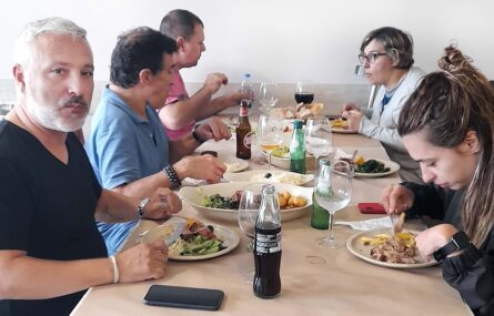 Cinco pessoas sentadas à mesa. Em cima da mesa pratos e travessas com comida, copos e garrafas.