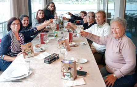 Fotografia com nove pessoas sentadas à mesa. Estão todas sorridentes e parecem estar a fazer um brinde usando, para tal, as chávenas de café.