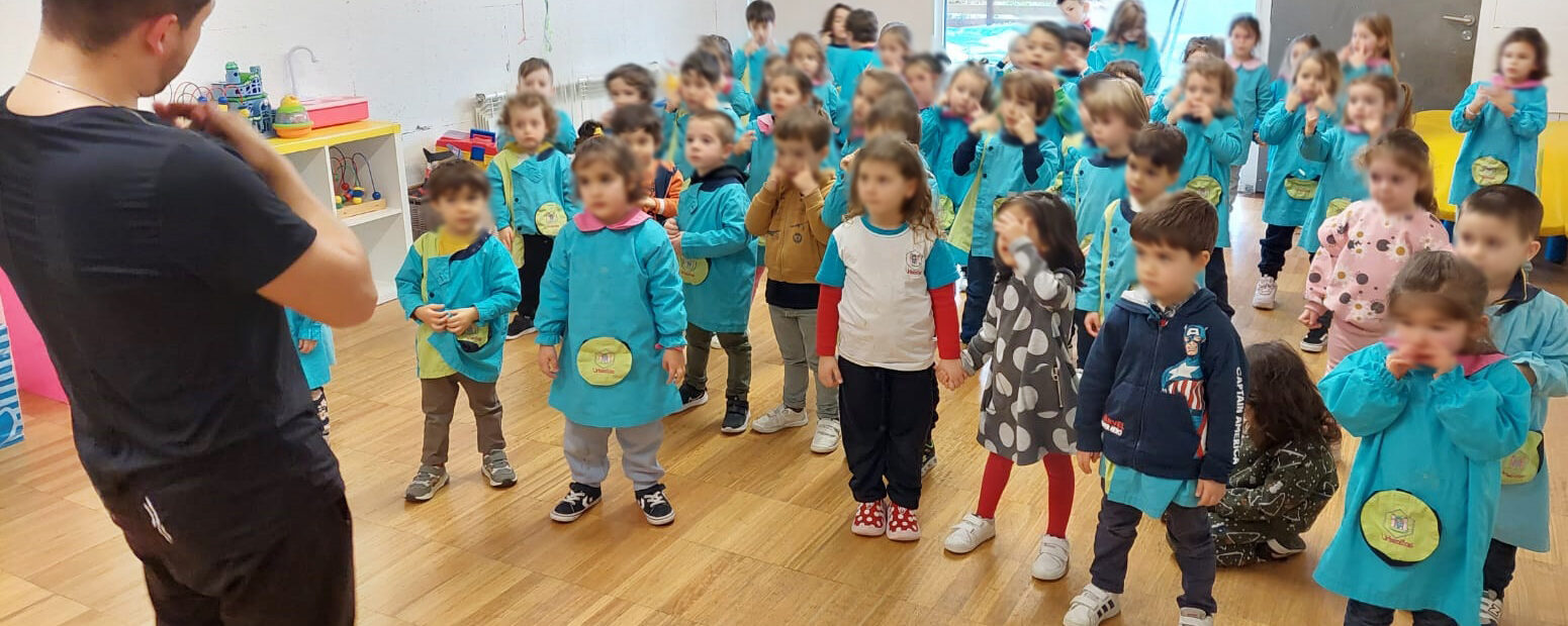 Em sala interior grupo de cerca de quatro dezenas de crianças acompanha explicação e gestos feitos por pessoa adulta. Os rostos das crianças foram todos desfocados.