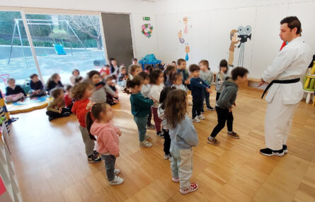 Em sala interior grupo de cerca de duas dezenas de crianças acompanha explicação e gestos feitos por pessoa adulta que fará uma apresentação de artes marciais. Algumas das crianças estão de pé, outras sentadas no chão, mais ao fundo da imagem. Os rostos das crianças foram todos desfocados.