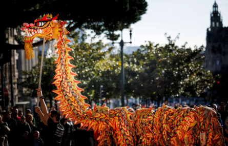 Fotografia de um dragão (típico das comemorações do Ano Novo Chinês) em tons laranja e amarelo. O dragão é elevado por várias pessoas. Ao fundo da imagem algumas árvores e pessoas na assistência.