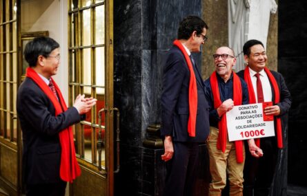 Quatro pessoas sorridentes, lado a lado. Todas usam um cachecol vermelho. Duas seguram um cartaz que indica “donativo para as associações de solidariedade”.